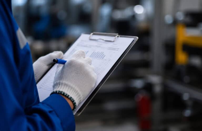 Factory technician wearing protective gloves recording inspection results on a clipboard during industrial equipment maintenance.