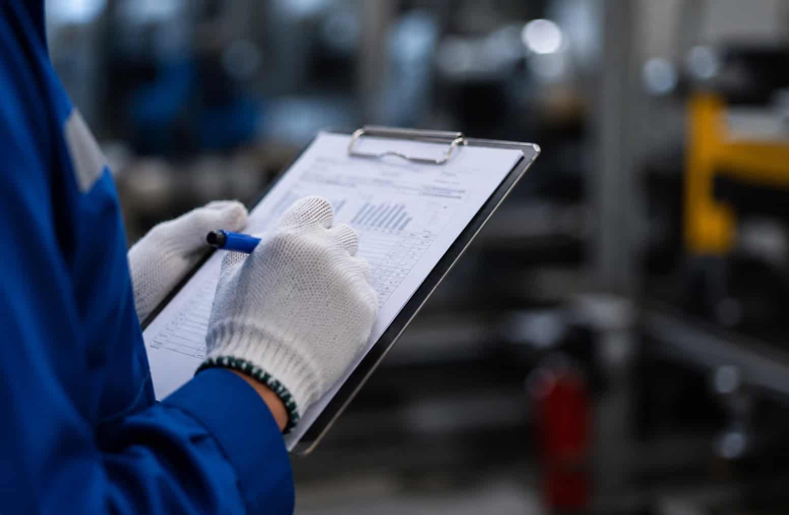 Factory technician wearing protective gloves recording inspection results on a clipboard during industrial equipment maintenance.