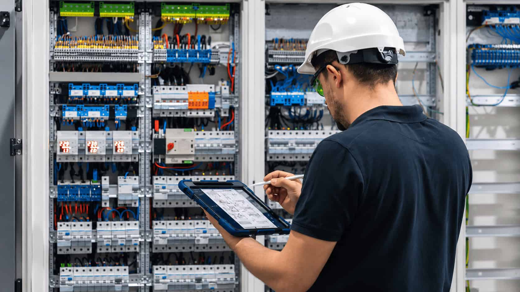 Industrial technician inspecting an electrical control panel with a rugged tablet during factory equipment maintenance.
