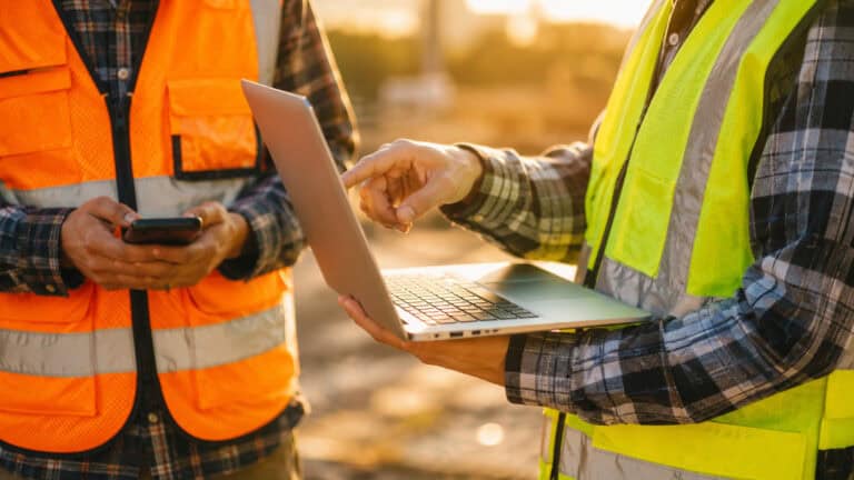 Field engineers wearing high-visibility safety vests using a laptop and mobile device on a construction site, representing remote monitoring, digital collaboration, and industrial IoT operations in an outdoor work environment.