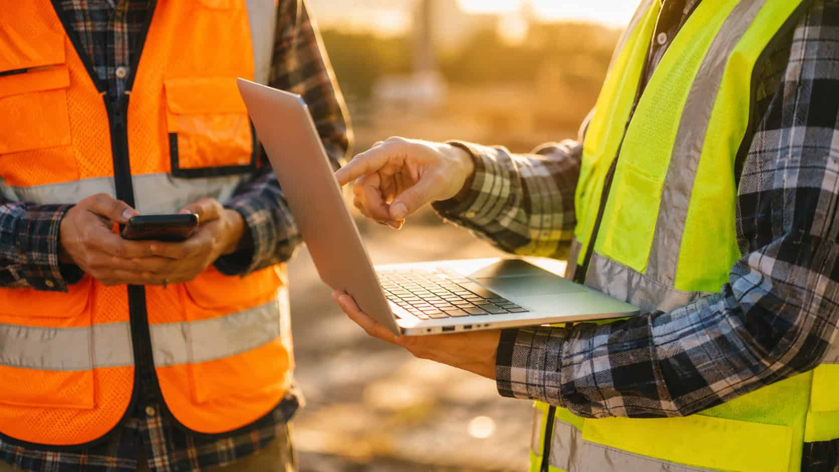 Field engineers wearing high-visibility safety vests using a laptop and mobile device on a construction site, representing remote monitoring, digital collaboration, and industrial IoT operations in an outdoor work environment.