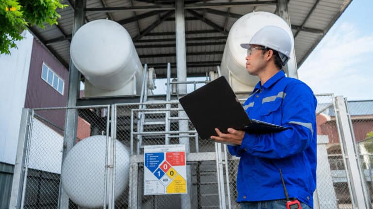 Industrial worker wearing a safety helmet and protective uniform inspecting equipment with a laptop at an outdoor industrial facility, with large storage tanks, safety barriers, and infrastructure in the background.