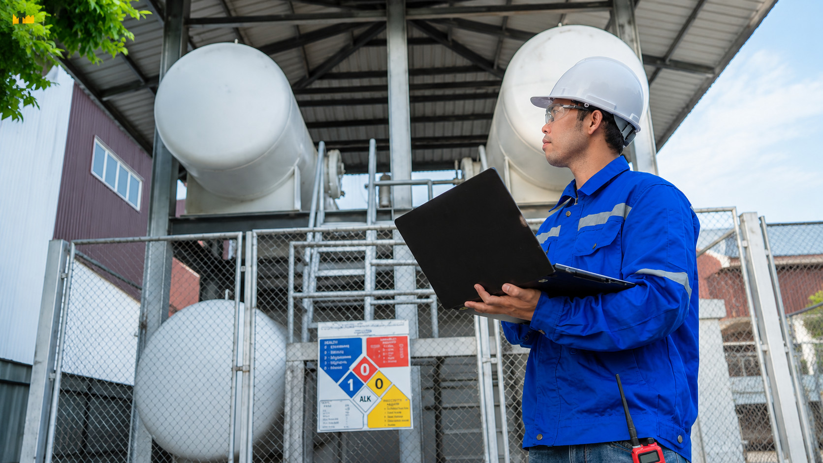 Industrial worker wearing a safety helmet and protective uniform inspecting equipment with a laptop at an outdoor industrial facility, with large storage tanks, safety barriers, and infrastructure in the background.
