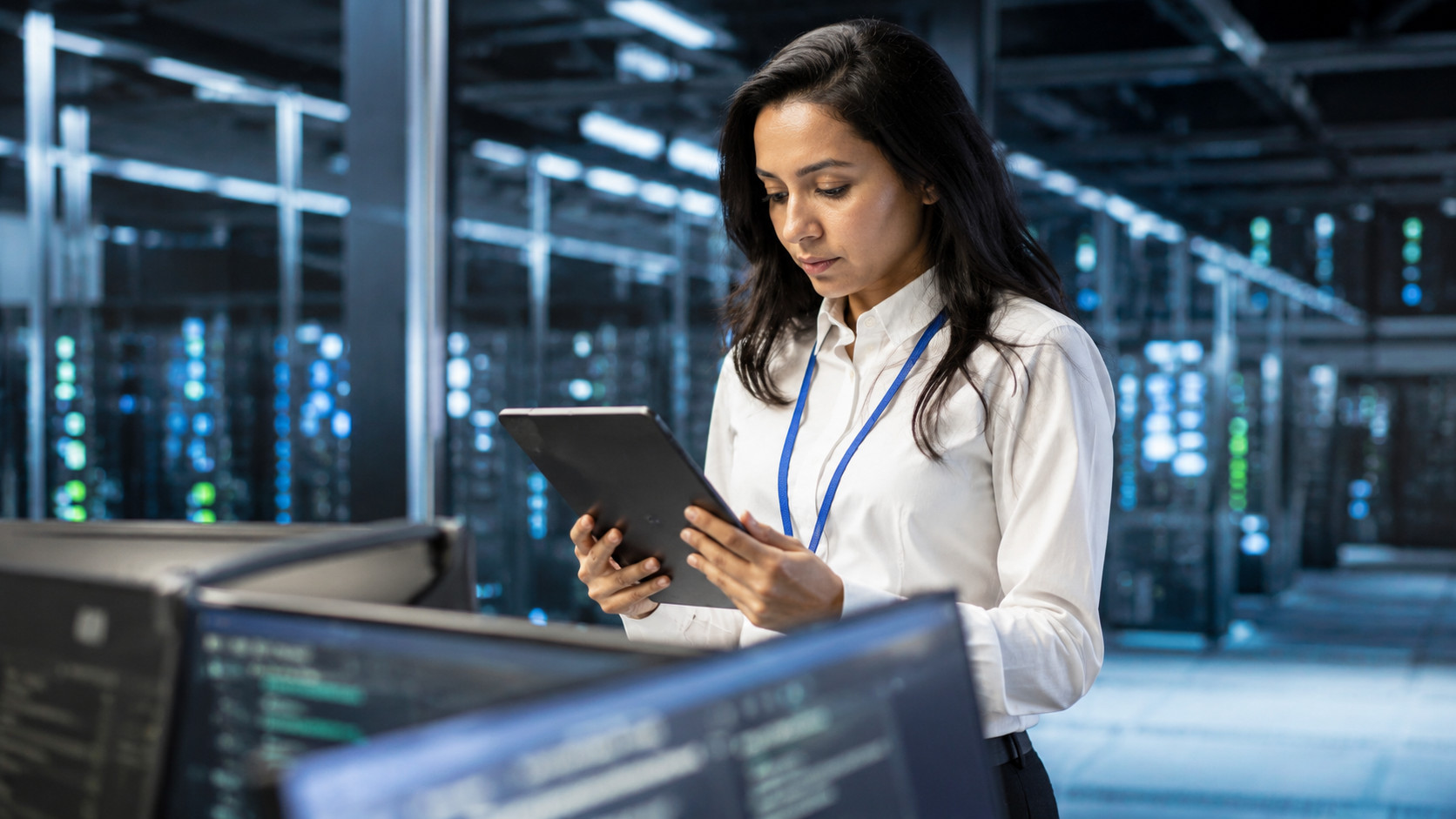 Professional data center engineer using a tablet while monitoring digital infrastructure, with server racks, technology equipment, and computer displays in a modern high-tech operations environment.