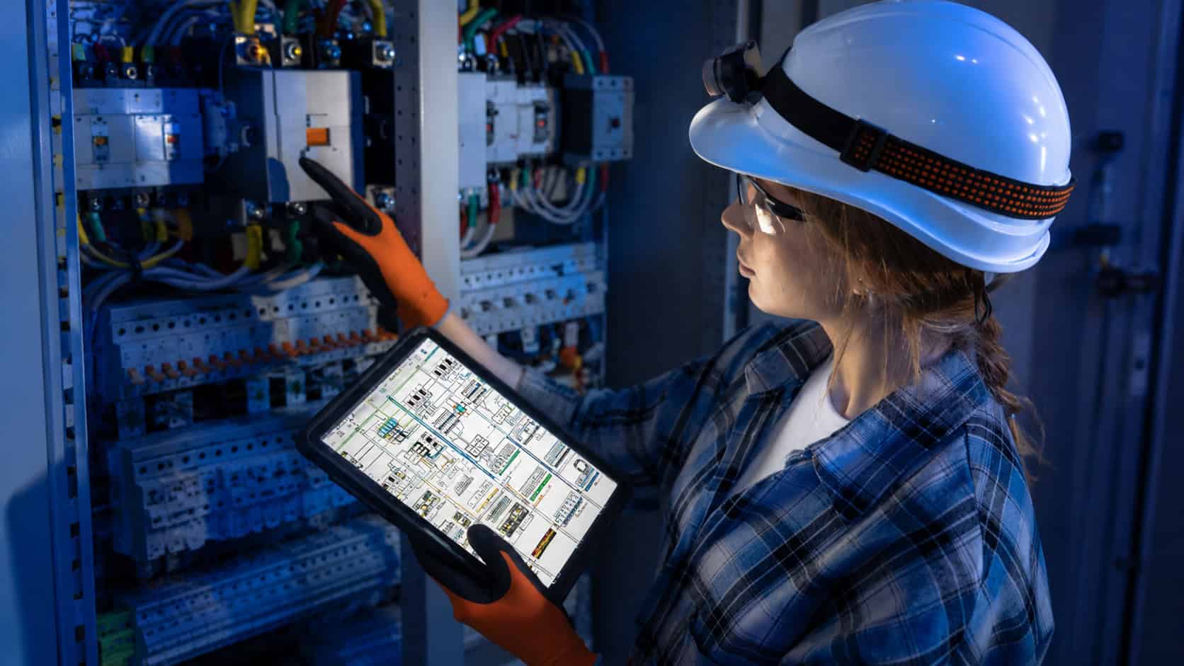 Engineer inspecting an industrial electrical control cabinet while using a tablet to monitor equipment status and wiring in a smart manufacturing facility.