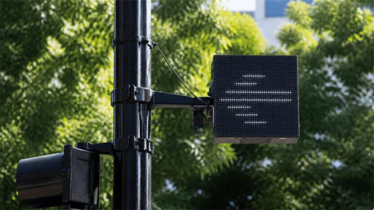 A black metal street pole supports a square LED matrix display with abstract white dots, set against sunlit green trees.
