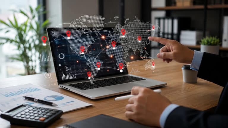 Business professional interacting with a digital world map showing connected global locations, data links, and analytics dashboards on a laptop in a modern office workspace.