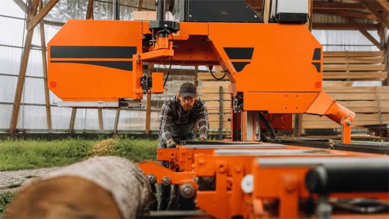 A worker wearing hearing protection operates a large bright orange sawmill machine in a timber-processing workshop.