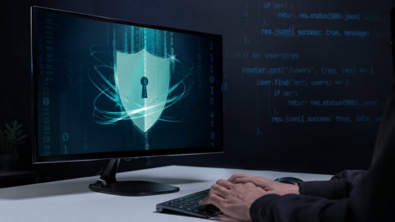 Computer monitor displaying a digital security shield interface with binary code and programming elements, while a user works at a desktop workstation, representing cybersecurity, data protection, and secure digital infrastructure.