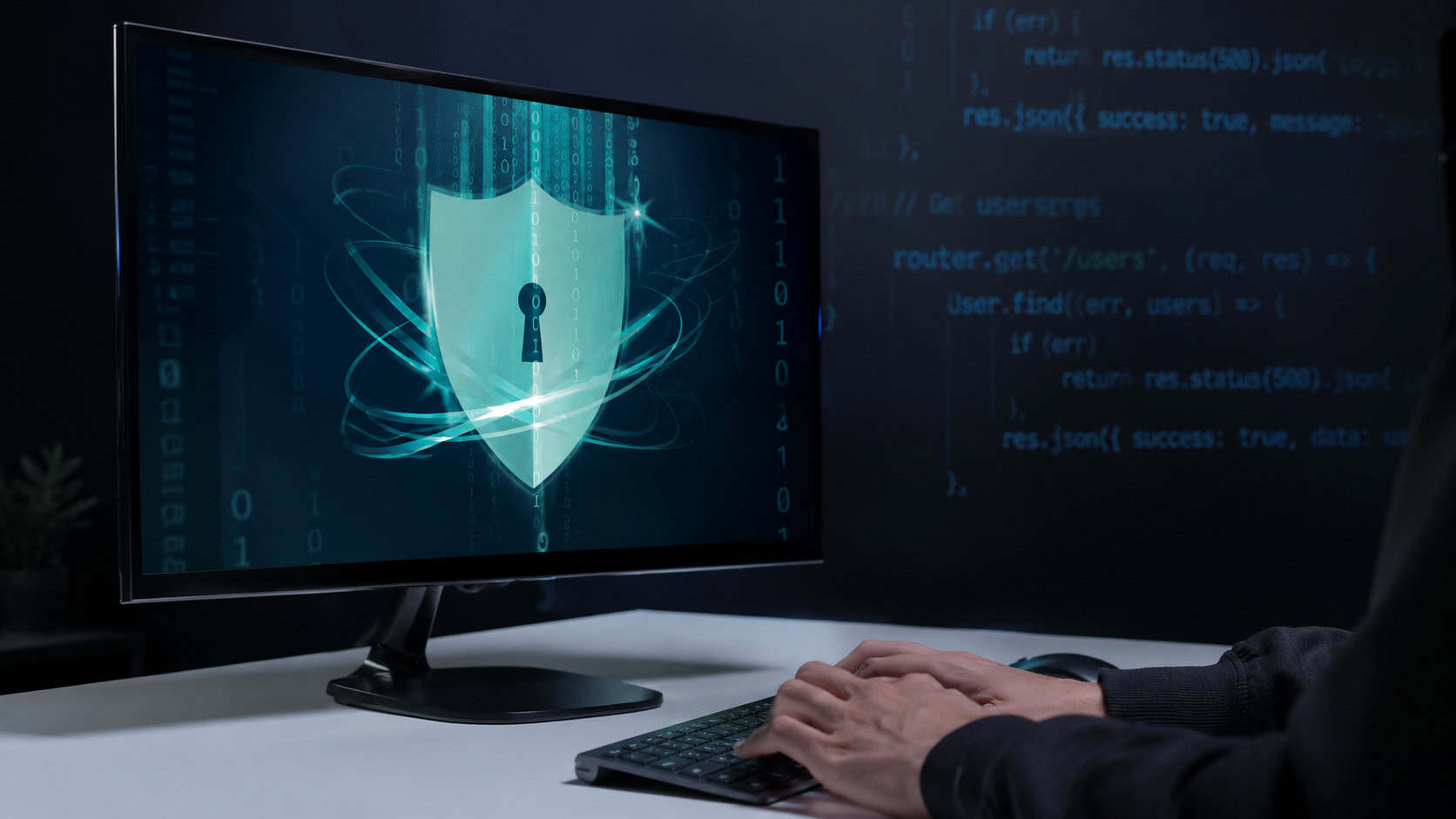 Computer monitor displaying a digital security shield interface with binary code and programming elements, while a user works at a desktop workstation, representing cybersecurity, data protection, and secure digital infrastructure.