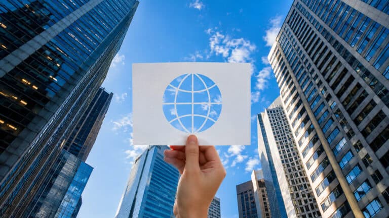 Hand holding a card with a global connectivity symbol against a modern city skyline, surrounded by high-rise buildings and blue sky, representing global communication networks, digital connectivity, and smart city infrastructure.