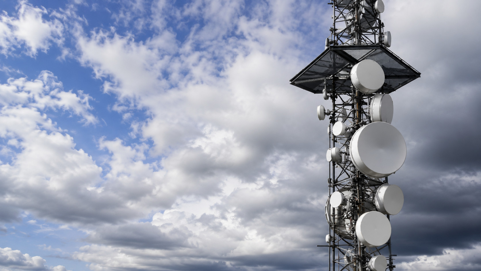 Telecommunication tower with multiple satellite dishes and wireless antennas against a cloudy sky, representing cellular networks, wireless communication infrastructure, and long-range connectivity.
