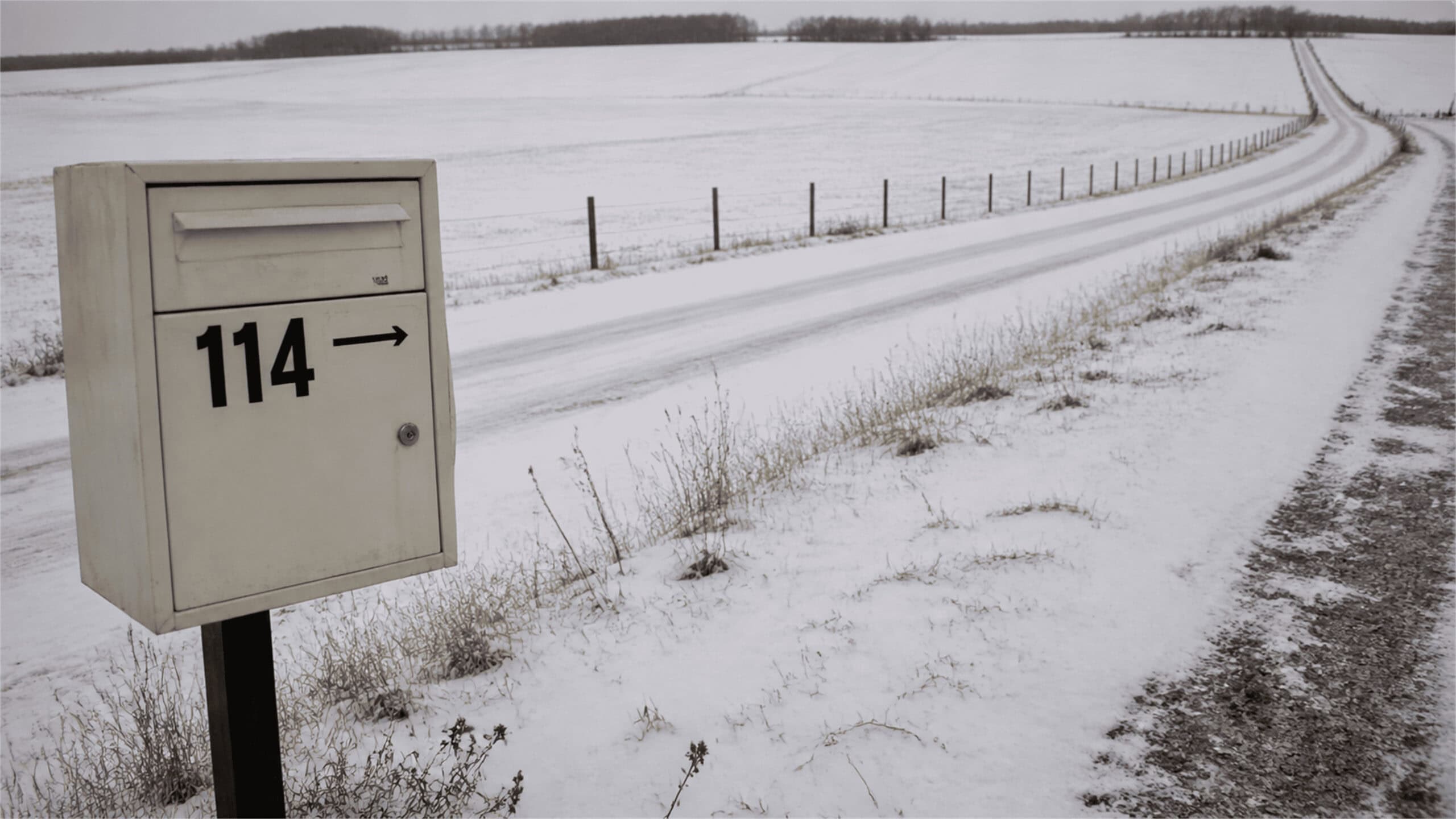 A weathered white mailbox marked 114 with a right arrow stands beside a snowy rural road winding past fenced winter fields.