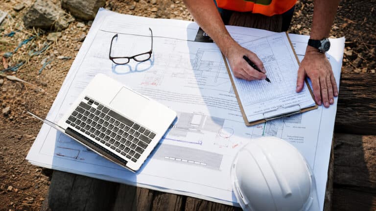 Overhead view of a construction engineer reviewing technical blueprints and inspection records at an outdoor worksite, with a laptop, safety helmet, glasses, and project documentation arranged on a wooden surface.