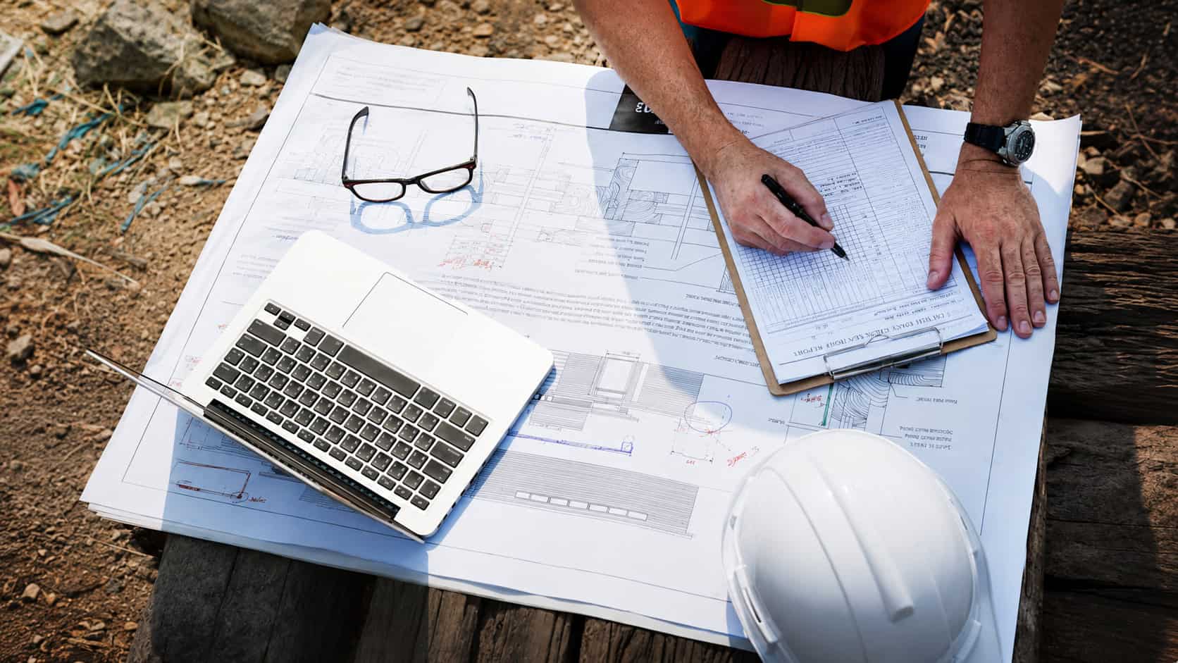 Overhead view of a construction engineer reviewing technical blueprints and inspection records at an outdoor worksite, with a laptop, safety helmet, glasses, and project documentation arranged on a wooden surface.