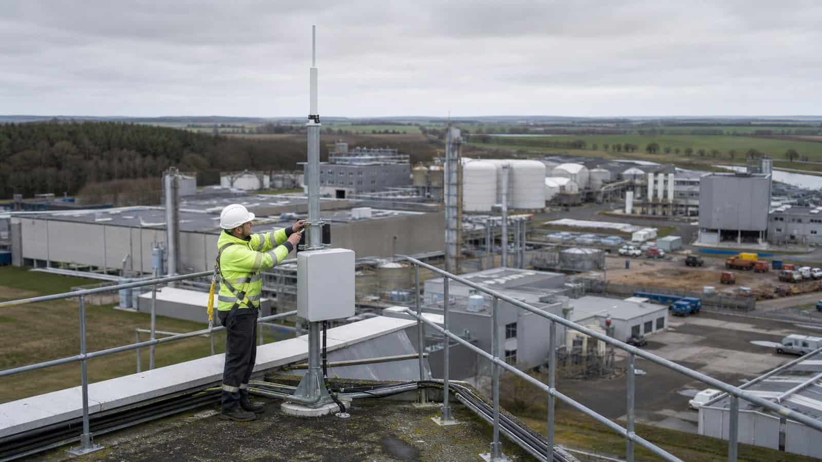 A rooftop technician checks an antenna mast overlooking an industrial campus, utility buildings and nearby fields.