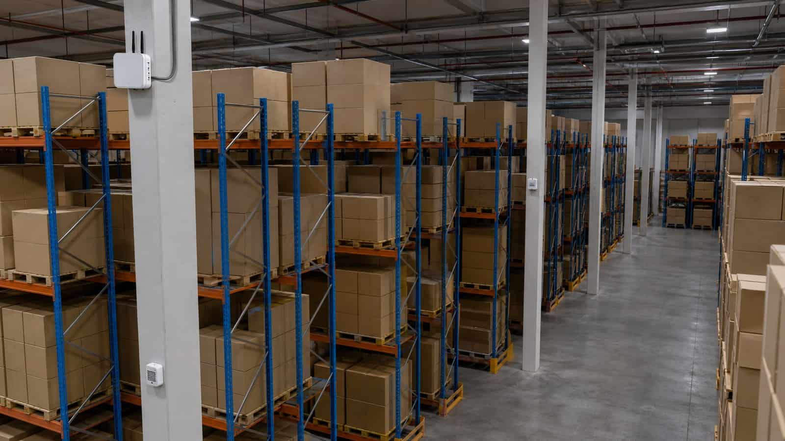 A warehouse aisle shows plain pallet racks, environmental sensors and a high-mounted gateway for capacity planning.