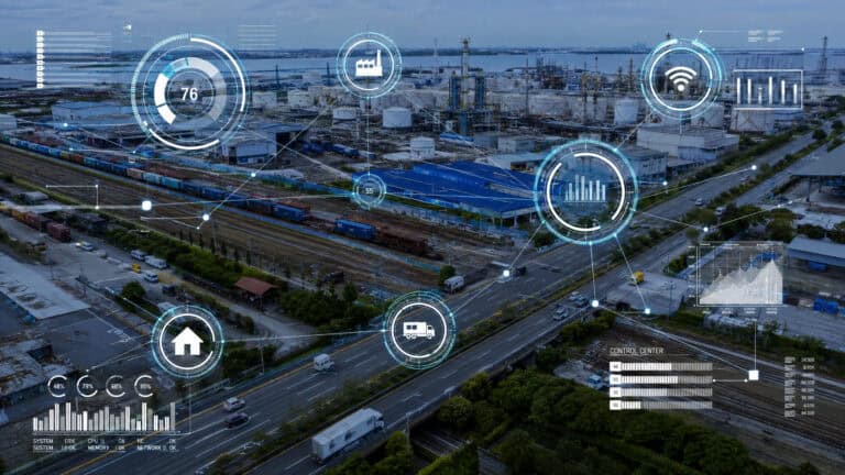 Aerial view of a large industrial facility and logistics hub with warehouses, storage tanks, railways, and highways, overlaid with futuristic digital HUD graphics showing connected systems, data analytics, IoT monitoring, and smart infrastructure management.