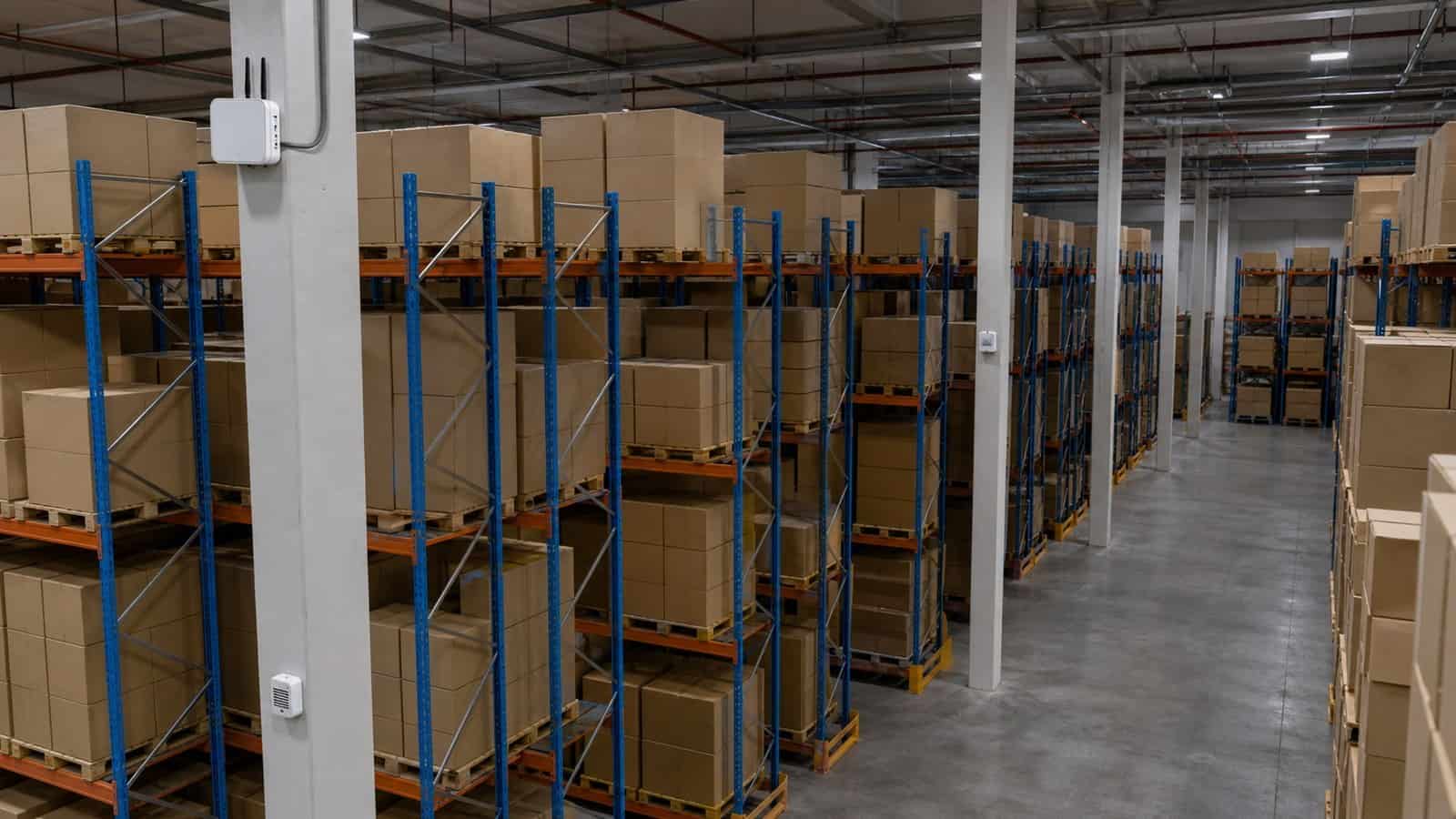 A warehouse aisle shows plain pallet racks, environmental sensors and a high-mounted gateway for capacity planning.