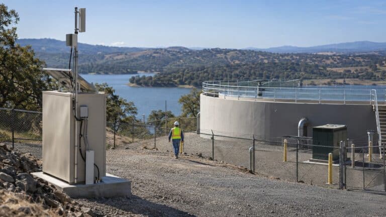 Field technician walks through a remote reservoir compound past telemetry, solar backup and cellular antenna equipment.